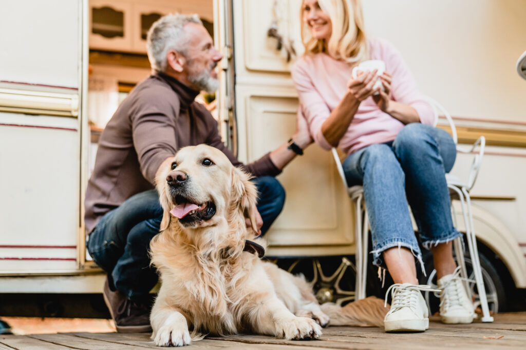 A senior couple sits outside their camper, making it feel like home. The man with gray hair and a beard kneels to pet their Golden Retriever, while the woman with blonde hair smiles, sitting comfortably on a chair with a cup in hand. The dog lies contentedly on the wooden floor.