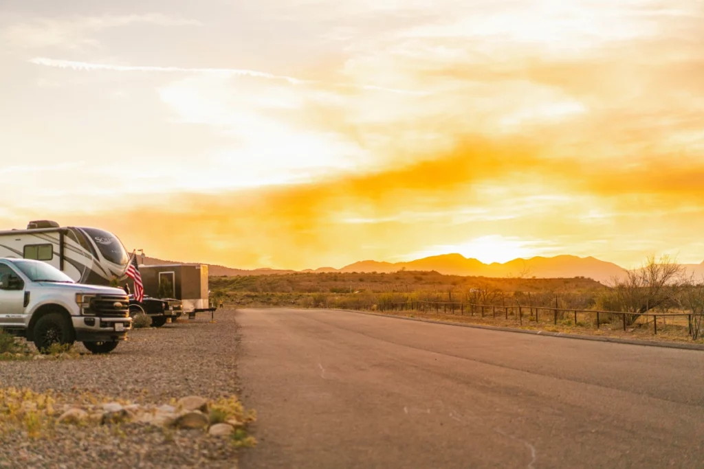 RVs and a truck parked on the side of a road in a desert landscape during sunset. The sky is orange and yellow, casting a warm glow over distant mountains and sparse vegetation.