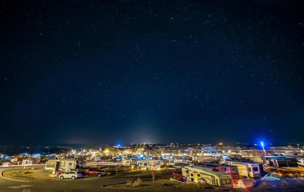 A nighttime scene at a trailer park with numerous RVs and trailers parked under a starry sky. The park is illuminated by streetlights and distant glowing city lights, creating a warm, inviting atmosphere against the vast, dark sky.
