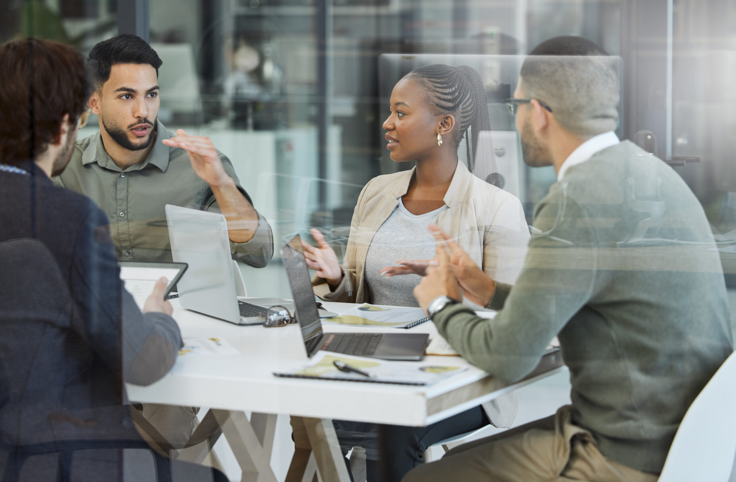 Four people sit around a table in a modern office, having a discussion. Laptops and documents are on the table, and two people gesture as they talk. The group appears to be collaborating or brainstorming.