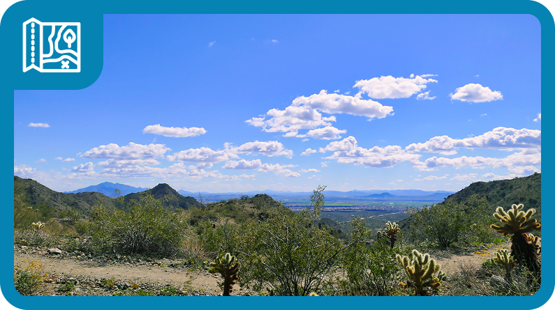 A scenic view of a desert landscape with mountains, shrubs, and cacti under a bright blue sky with scattered clouds. A turquoise border frames the image with a map icon in the top left corner.