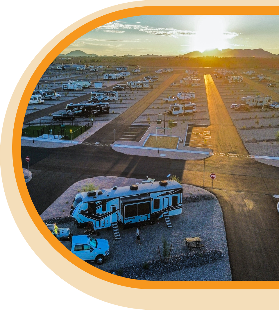 Aerial view of an RV park at sunset, with rows of RVs and trailers parked on gravel lots along paved roads, and mountains visible in the distance.