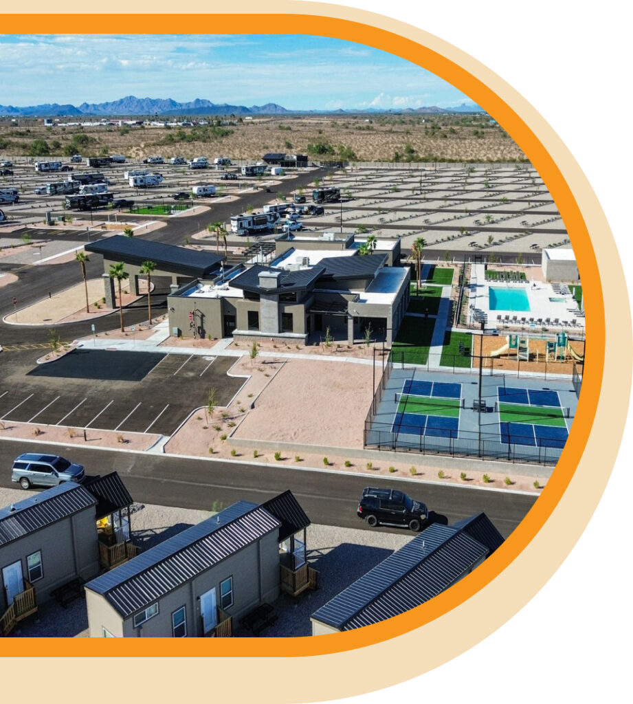 Aerial view of a modern RV resort with roads, parked RVs, small cabins, a central clubhouse, swimming pool, and pickleball courts in a desert landscape with mountains in the distance.