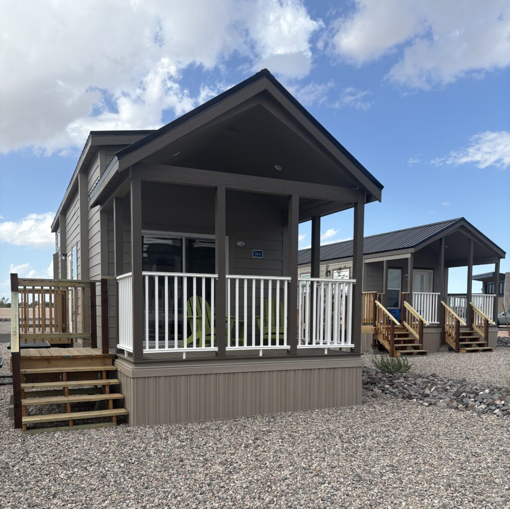 A small, modern tiny house with a covered porch, white railings, and steps leading up, set on a gravel lot under a partly cloudy sky. Another similar tiny house is visible in the background.