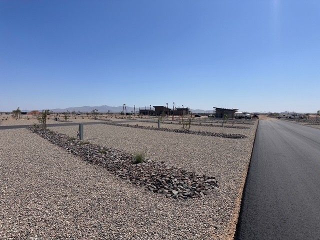 A newly paved road runs through a desert landscape with gravel lots and sparse plants. Buildings are visible in the distance under a clear blue sky, with mountains on the horizon.