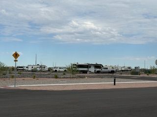 Several RVs and trailers are parked in a gravel lot with desert vegetation under a partly cloudy sky. A yellow road sign is visible on the left, and a paved road runs in the foreground.