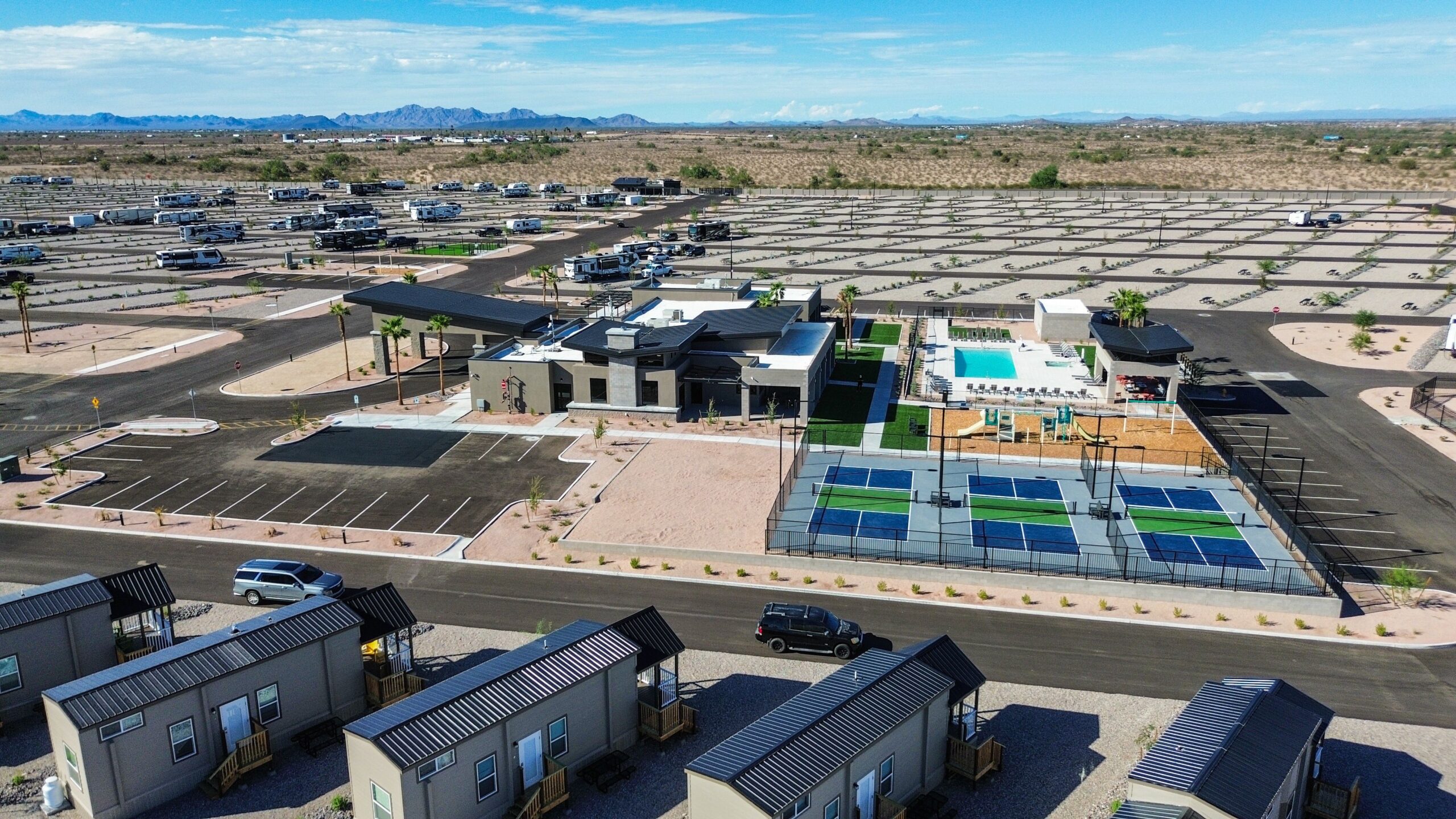 Aerial view of an RV park with rows of RV parking spaces, several small cabins, a central building, swimming pool, and pickleball courts, surrounded by desert landscape and mountains in the distance.
