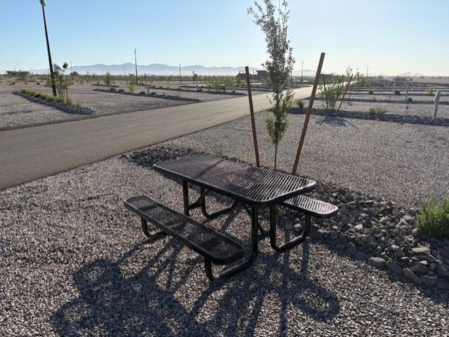 A metal picnic table with two benches sits on gravel in an empty, sparsely landscaped RV park under a clear sky, with mountains visible in the distant background.