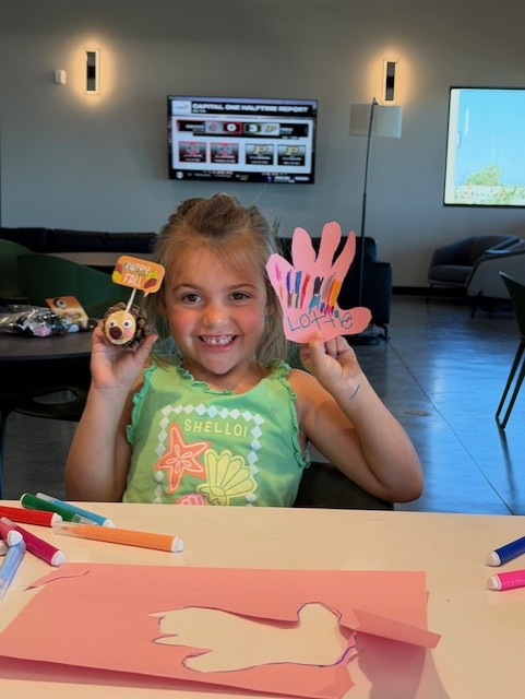 A smiling young girl sits at a table holding a handmade turkey craft and a colorful paper hand with "Lottie" written on it. Markers and craft supplies are spread out in front of her in a bright room.