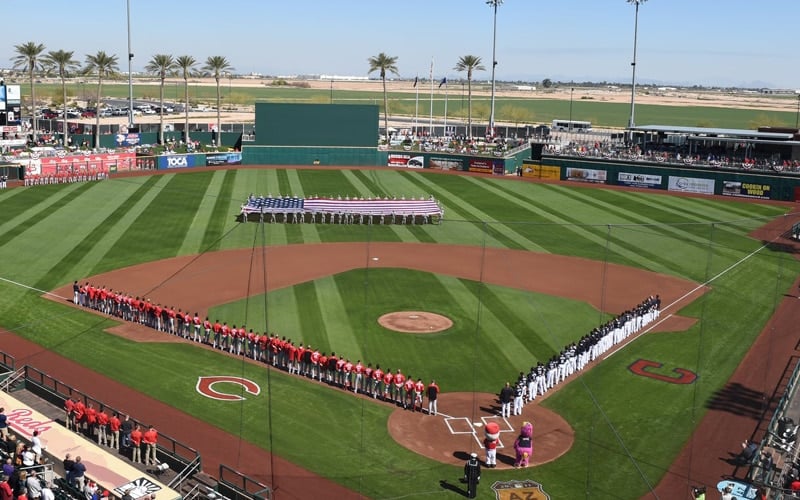 Baseball teams line up along the baselines for a pregame ceremony on a green field, with an American flag displayed in the outfield and palm trees and stadium seating in the background.