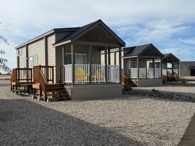 Three small, modern cabins with porches and yellow chairs are lined up on a gravel lot under a partly cloudy sky. Each cabin has steps leading up to the porch and white railings.
