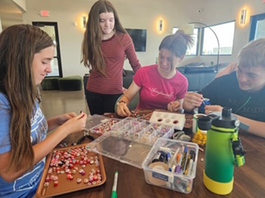 Four teens sit and stand around a table making jewelry with colorful beads and craft supplies. Smiling and focused, they work together in a bright room with large windows and modern decor.