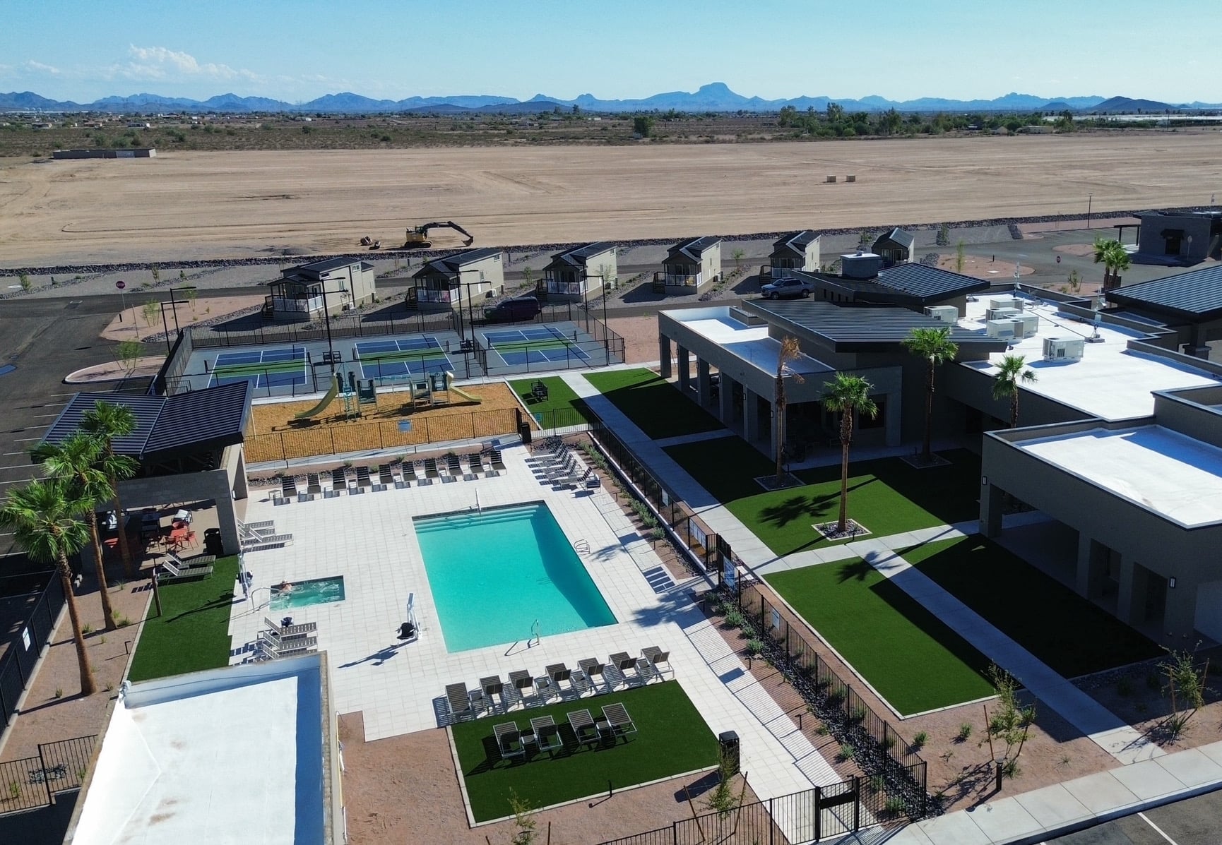 Aerial view of a modern recreational facility with a rectangular swimming pool, lounge chairs, playground, green lawns, palm trees, and surrounding buildings; desert landscape and mountains in the background.