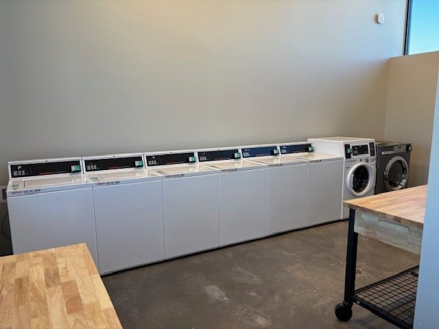 Six white washing machines and two dryers are lined up against a plain wall in a laundry room with a concrete floor and light wood tables.