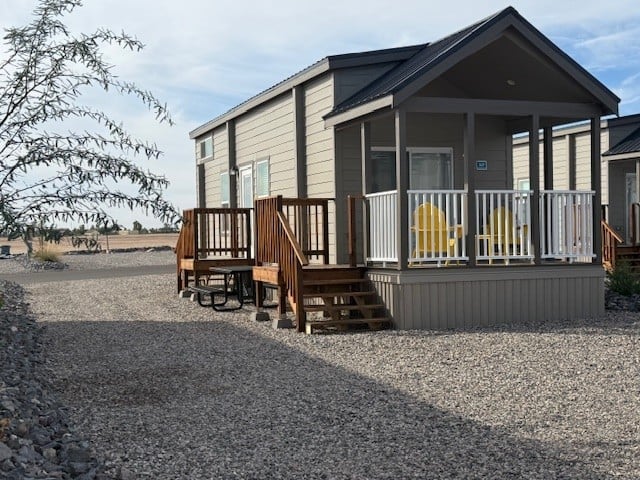 A small, modern cabin with a porch featuring two yellow chairs and wooden steps stands on a gravel lot. A picnic table sits nearby, and sparse vegetation is visible under a partly cloudy sky.