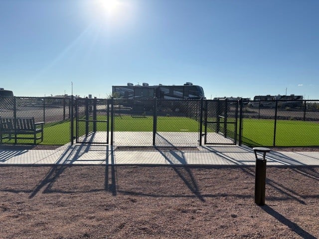 A fenced-in, grassy dog park area with gates, surrounded by pavement and gravel, sits in front of several parked RVs under a clear blue sky with the sun shining brightly.