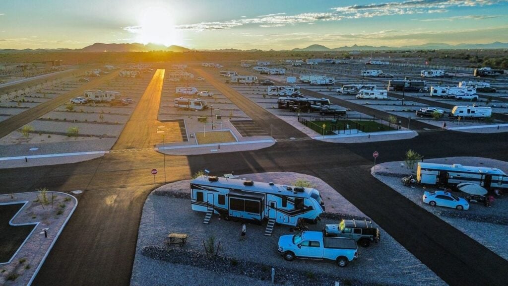 Aerial view of an RV park at sunset, showing multiple RVs and trailers parked in designated spots with paved roads, surrounded by desert landscape and distant mountains under a partly cloudy sky.