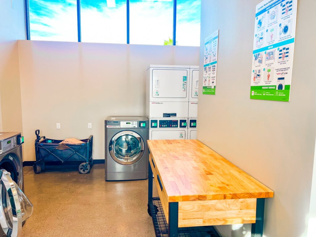 Laundry room with washers, dryers, a wooden folding table, a black laundry cart, and instructional posters on the wall. Large windows let in natural light.