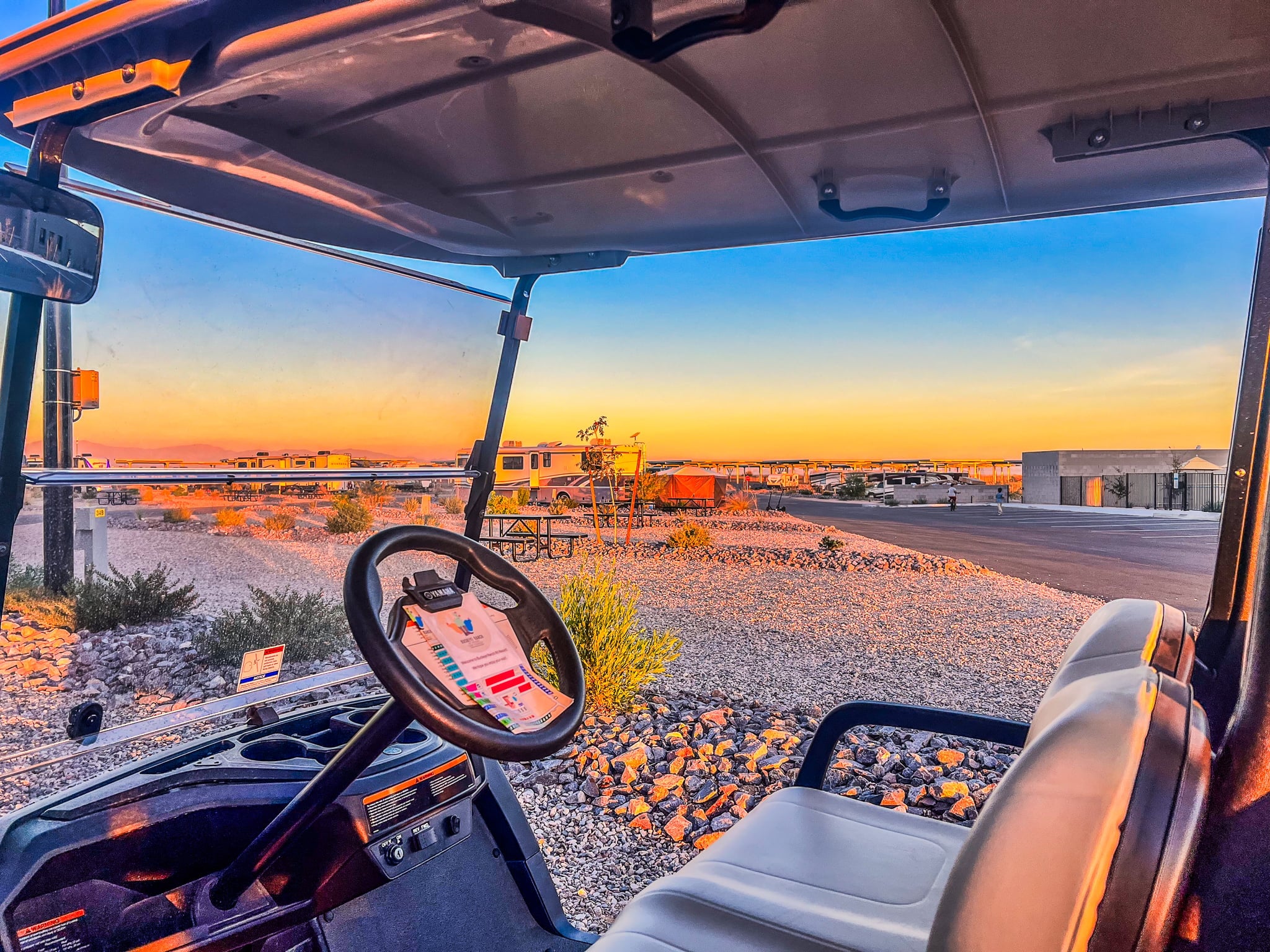 A golf cart with white seats parked on gravel near a picnic table, with an RV park and vibrant orange and blue sunset sky in the background.