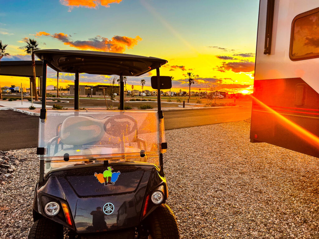A golf cart parked on gravel near an RV at sunset, with palm trees and other RVs in the background under a colorful sky with orange and yellow hues.