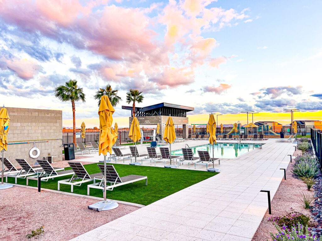 Outdoor swimming pool area with lounge chairs and closed yellow umbrellas, surrounded by palm trees and a fence, under a colorful sunset sky with scattered clouds.