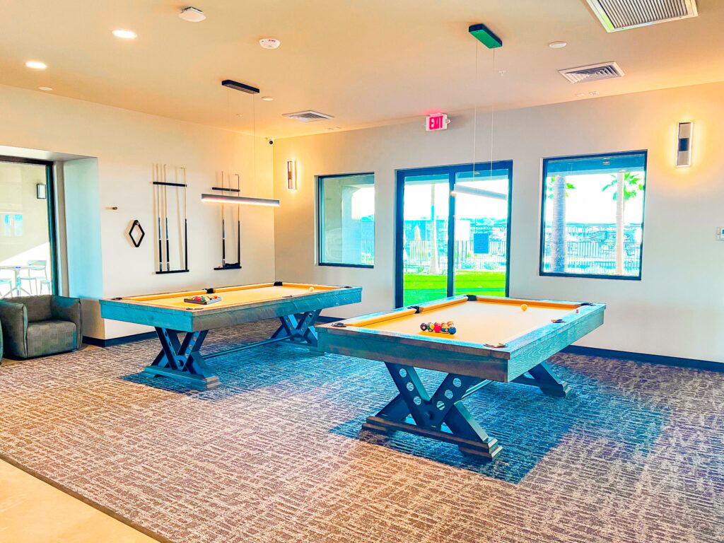 A bright game room with two pool tables, neatly arranged cues on the wall, large windows letting in natural light, and modern light fixtures hanging from the ceiling.