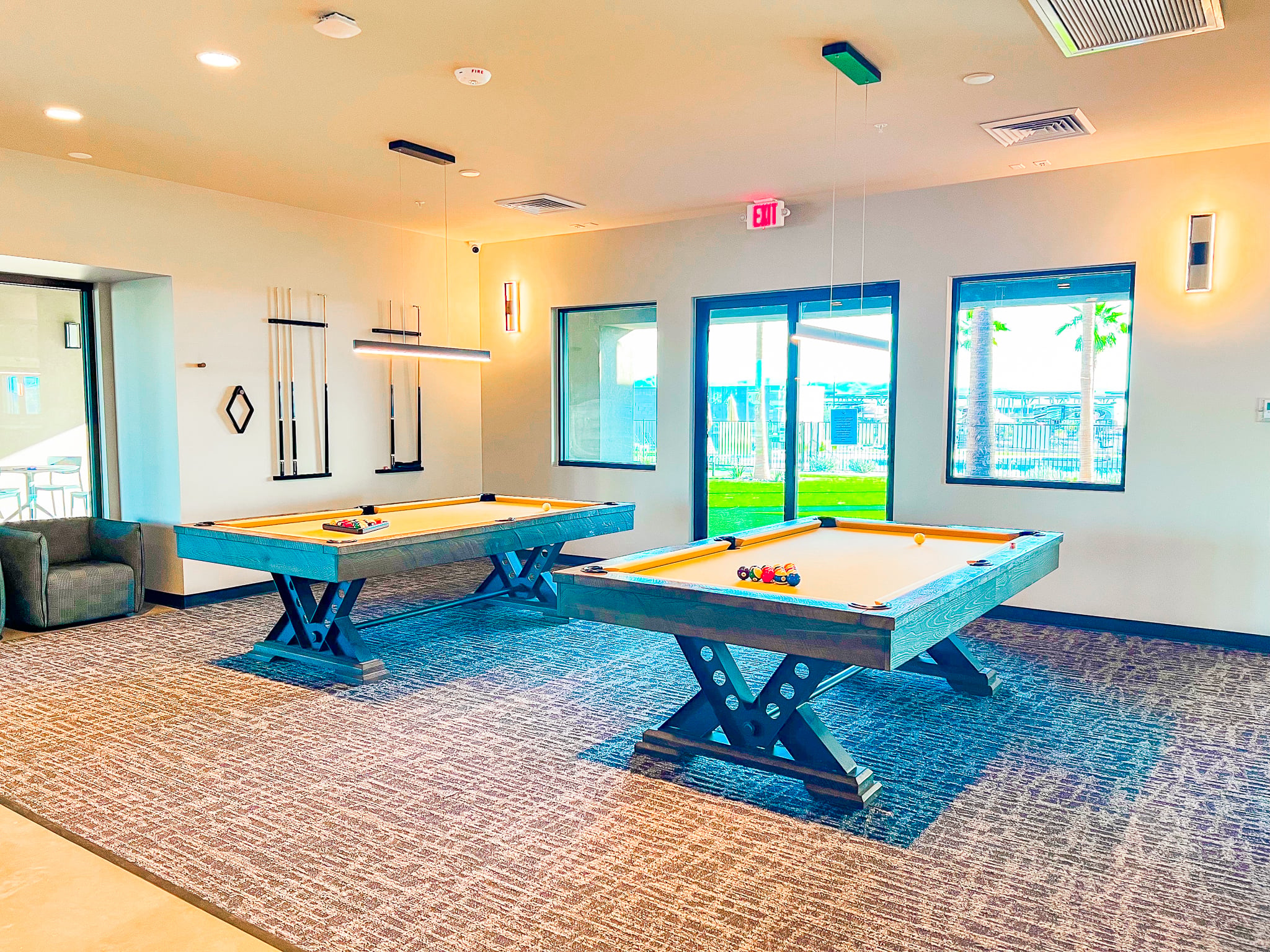 A bright game room with two pool tables, neatly arranged cues on the wall, large windows letting in natural light, and modern light fixtures hanging from the ceiling.