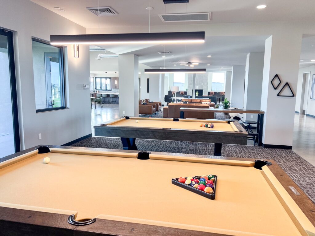 A modern game room with two pool tables, one set up for a game, and a lounge area with sofas and tables in the background. The space is bright with large windows and contemporary lighting.