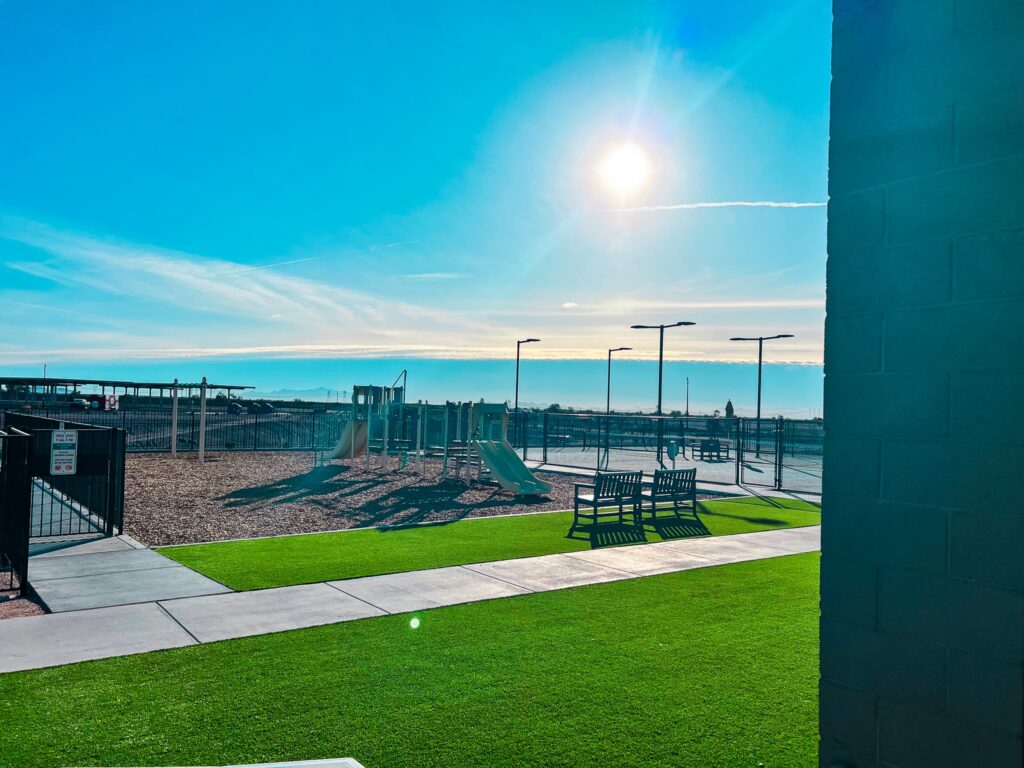 A sunny day at an empty playground with slides and climbing equipment, bordered by a fence. Benches sit on bright green artificial grass, and a building wall is visible on the right. The sun shines brightly in a clear blue sky.