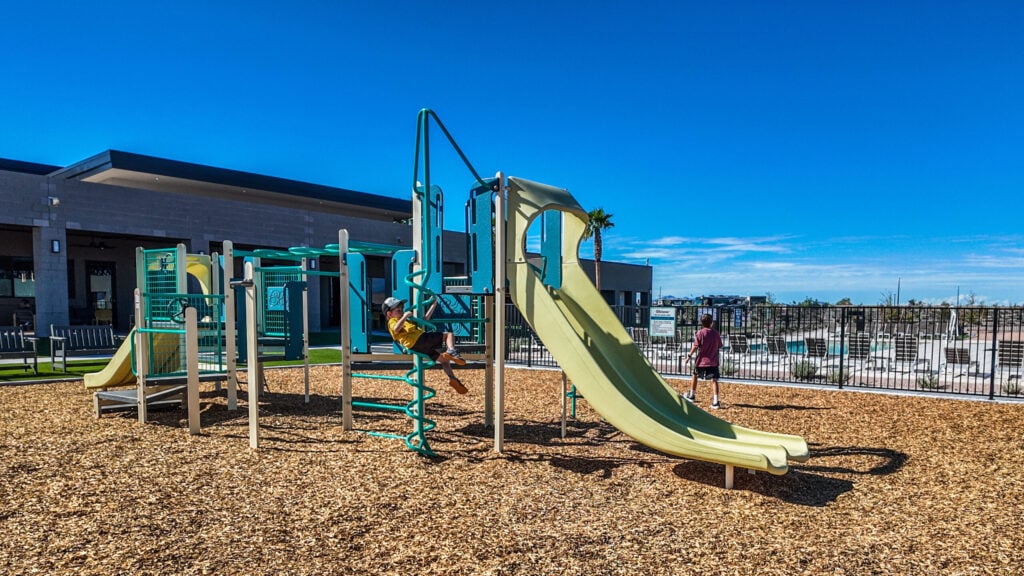 Children playing on a playground with slides, ladders, and climbing bars, surrounded by wood chips and a fence, under a clear blue sky near a modern building.