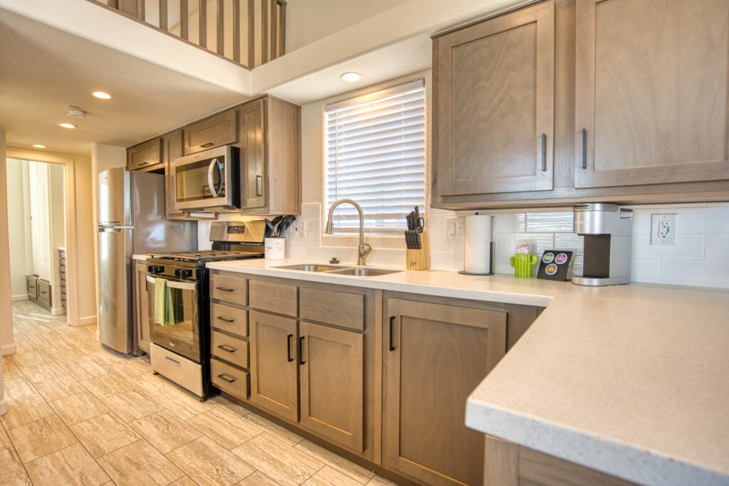 Modern kitchen with light wood cabinets, white countertops, stainless steel appliances, a window with blinds, and a set of knives and utensils on the counter. The floor has beige tiles and there is overhead lighting.