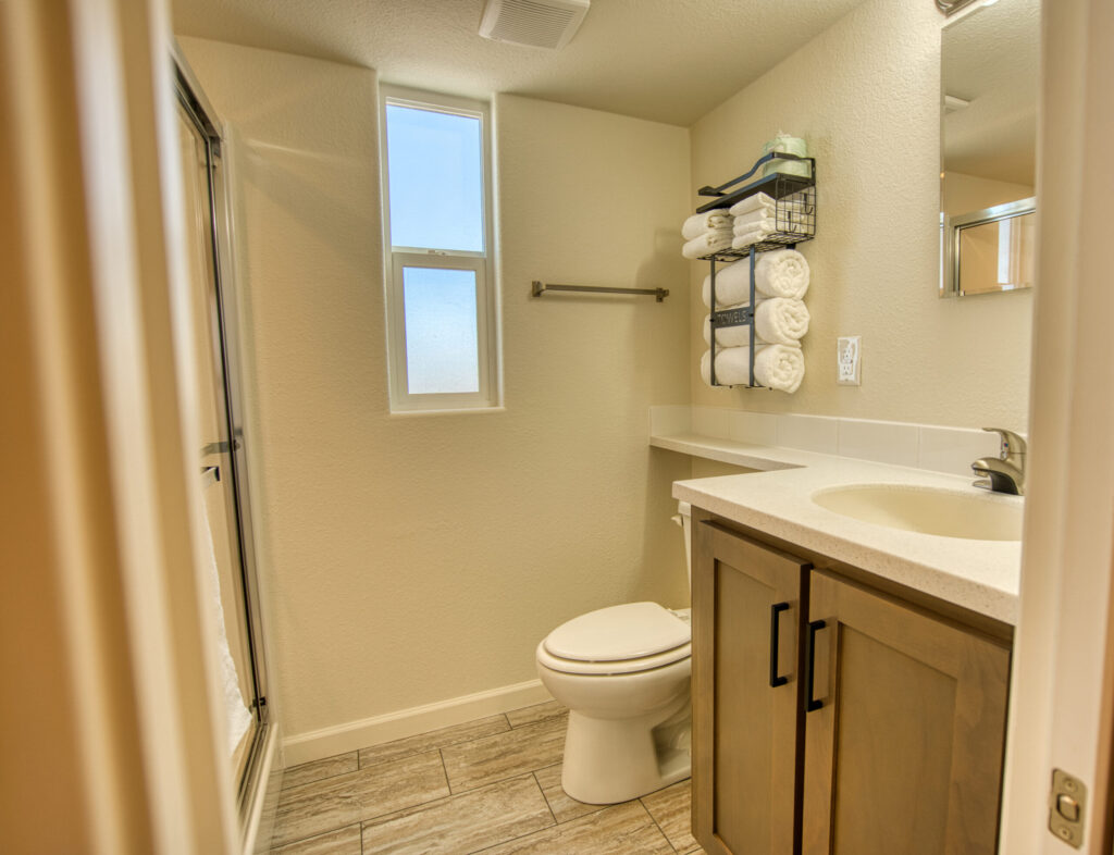 A clean, modern bathroom with wood cabinets, a white countertop, a toilet, a glass shower, a wall shelf with neatly rolled towels, and a frosted window letting in natural light.