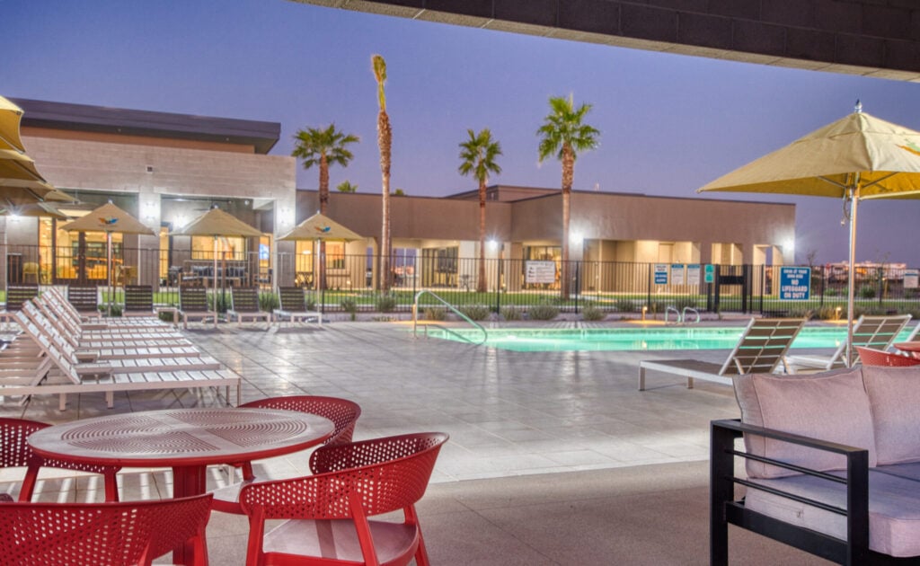 Outdoor pool area at dusk with lounge chairs, yellow umbrellas, and red patio chairs in the foreground. Palm trees and a modern building are visible in the background, illuminated by soft lighting.
