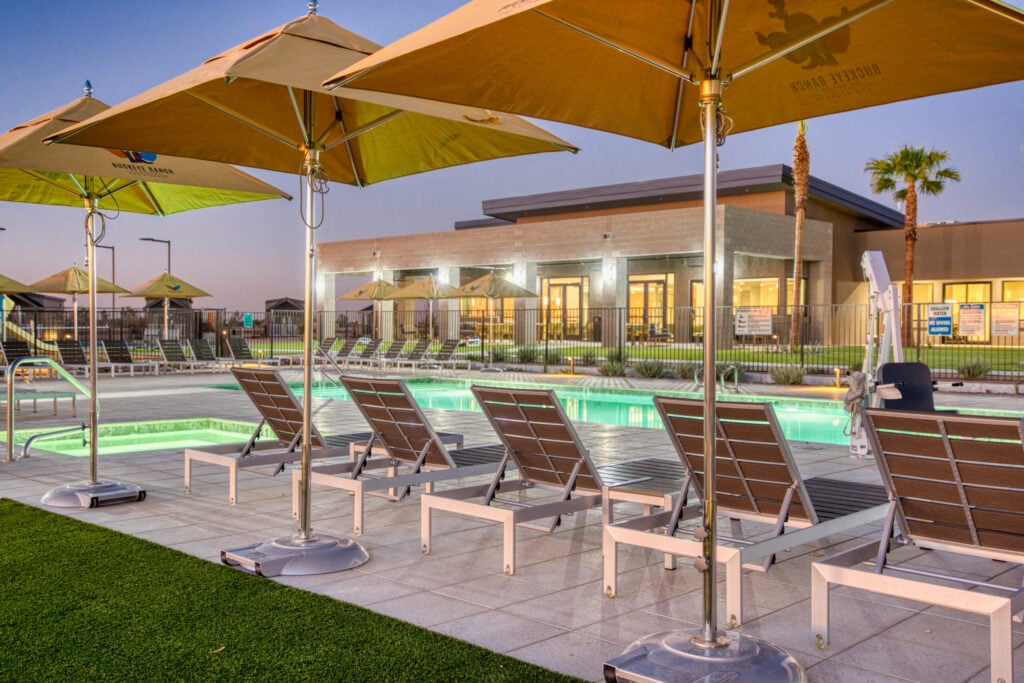 Poolside lounge chairs under large yellow umbrellas beside a modern building at sunset. The lit swimming pool is surrounded by palm trees, a fence, and manicured grass, creating a relaxing, inviting atmosphere.