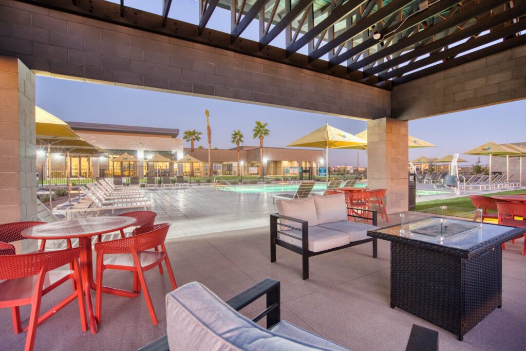 Covered patio with modern outdoor seating, red chairs, and tables overlooks a swimming pool area with lounge chairs, yellow umbrellas, and palm trees at dusk. Buildings and lights are visible in the background.