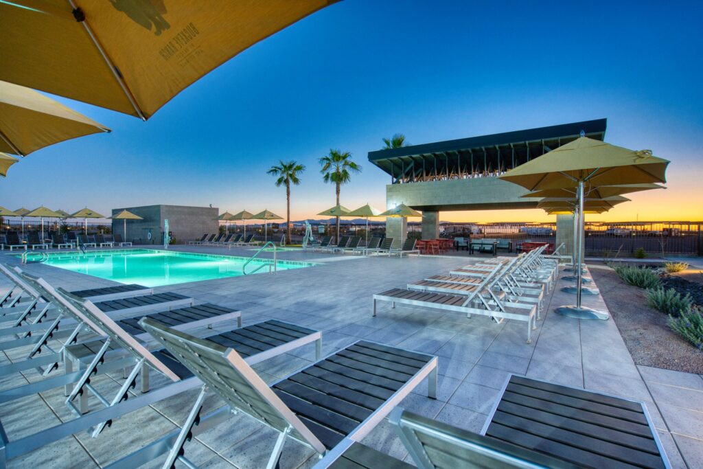 Modern outdoor pool area at sunset with lounge chairs, large yellow umbrellas, palm trees, and a contemporary building in the background. The pool is illuminated, creating a relaxing atmosphere.