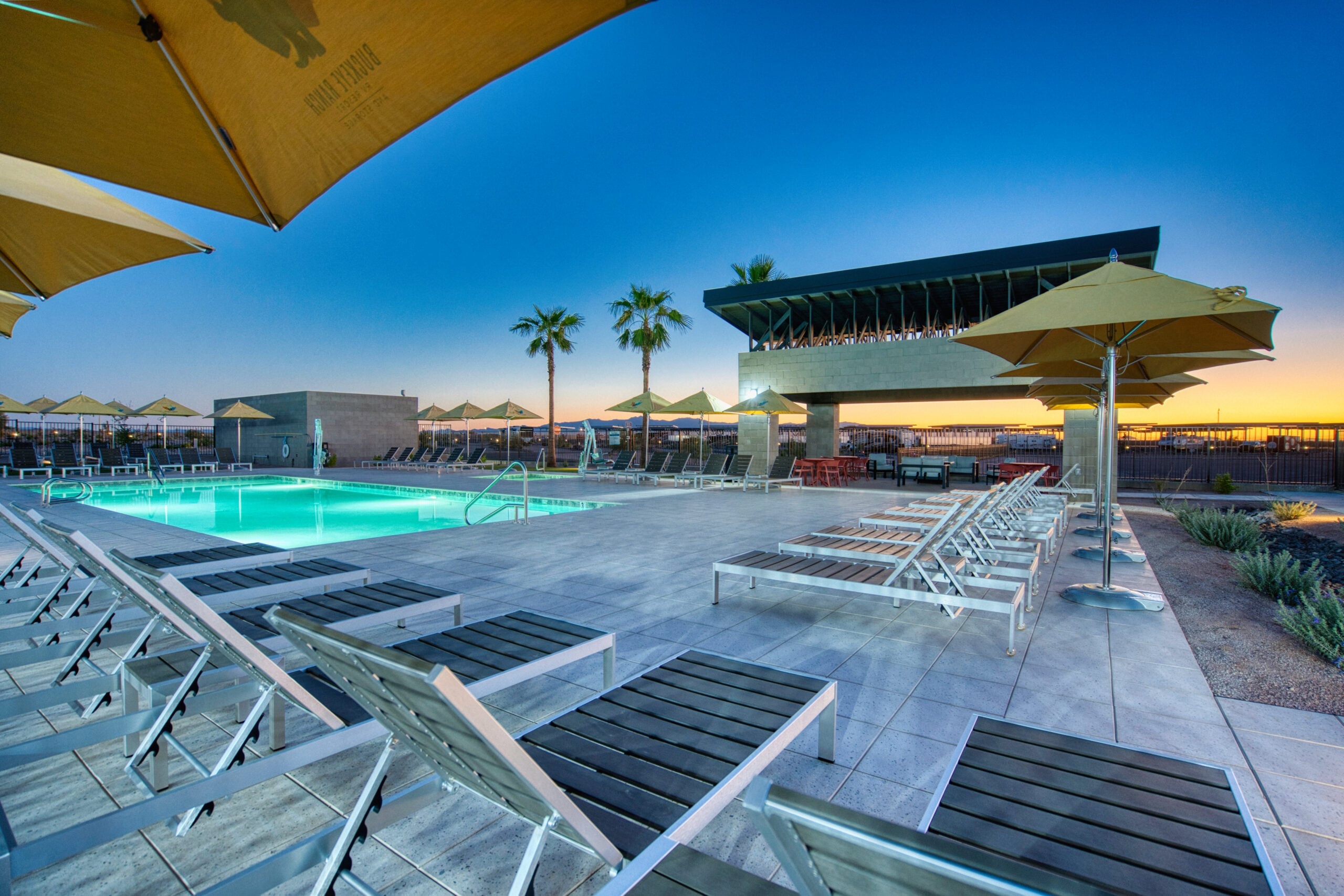 Outdoor pool area at sunset with lounge chairs, large yellow umbrellas, palm trees, and a modern building. The pool is illuminated, and the sky is clear, creating a relaxing, resort-like atmosphere.