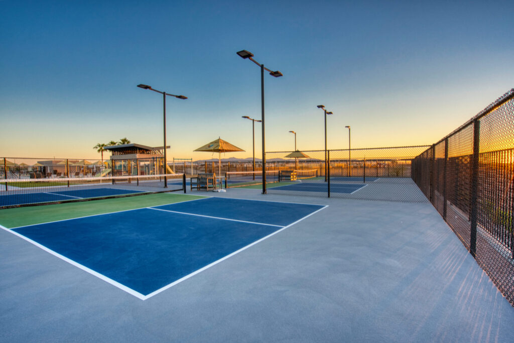 A modern outdoor tennis court complex at sunset, featuring several courts with blue and green surfaces, black chain-link fencing, tall light poles, and a shaded seating area in the background.