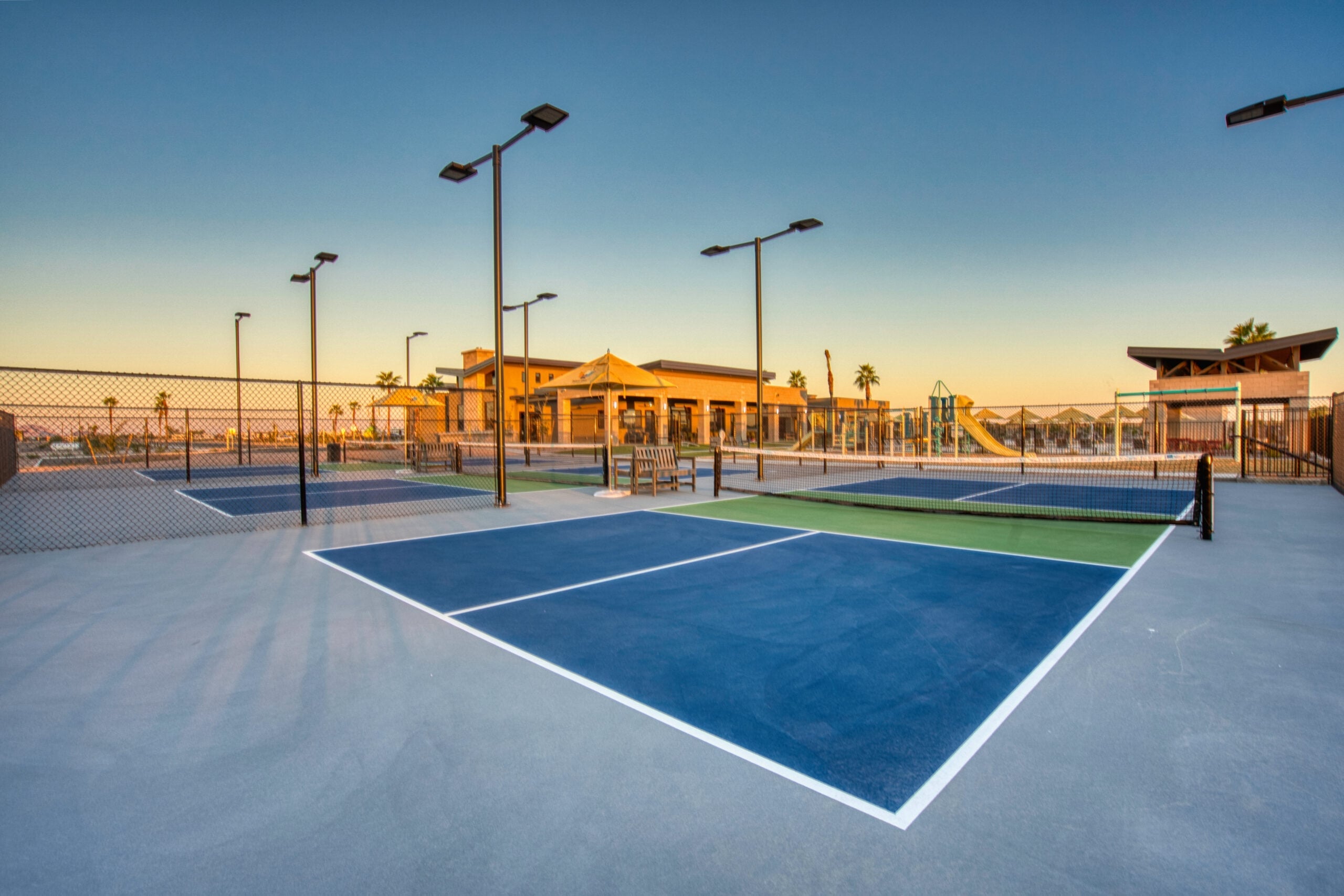 Empty outdoor pickleball courts with blue and green surfaces, surrounded by fencing and tall lights, near a modern building and a playground under a clear sky at sunset.