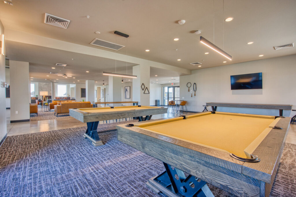 A modern game room with two pool tables, blue carpet, lounge seating in the background, wall-mounted TV, and minimalist decor in neutral tones. Large windows provide natural light.