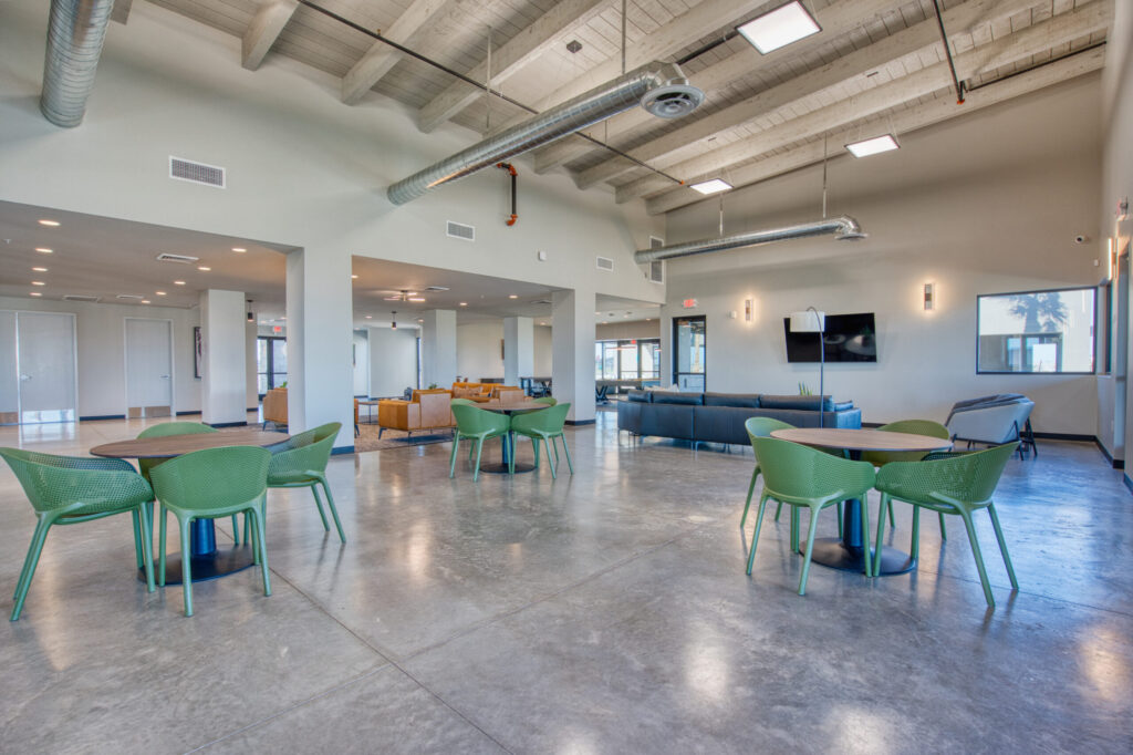 Modern, spacious common area with polished concrete floors, round tables with green chairs, blue sofas, wall-mounted TV, large windows, and exposed ceiling pipes. Natural light fills the open, airy space.