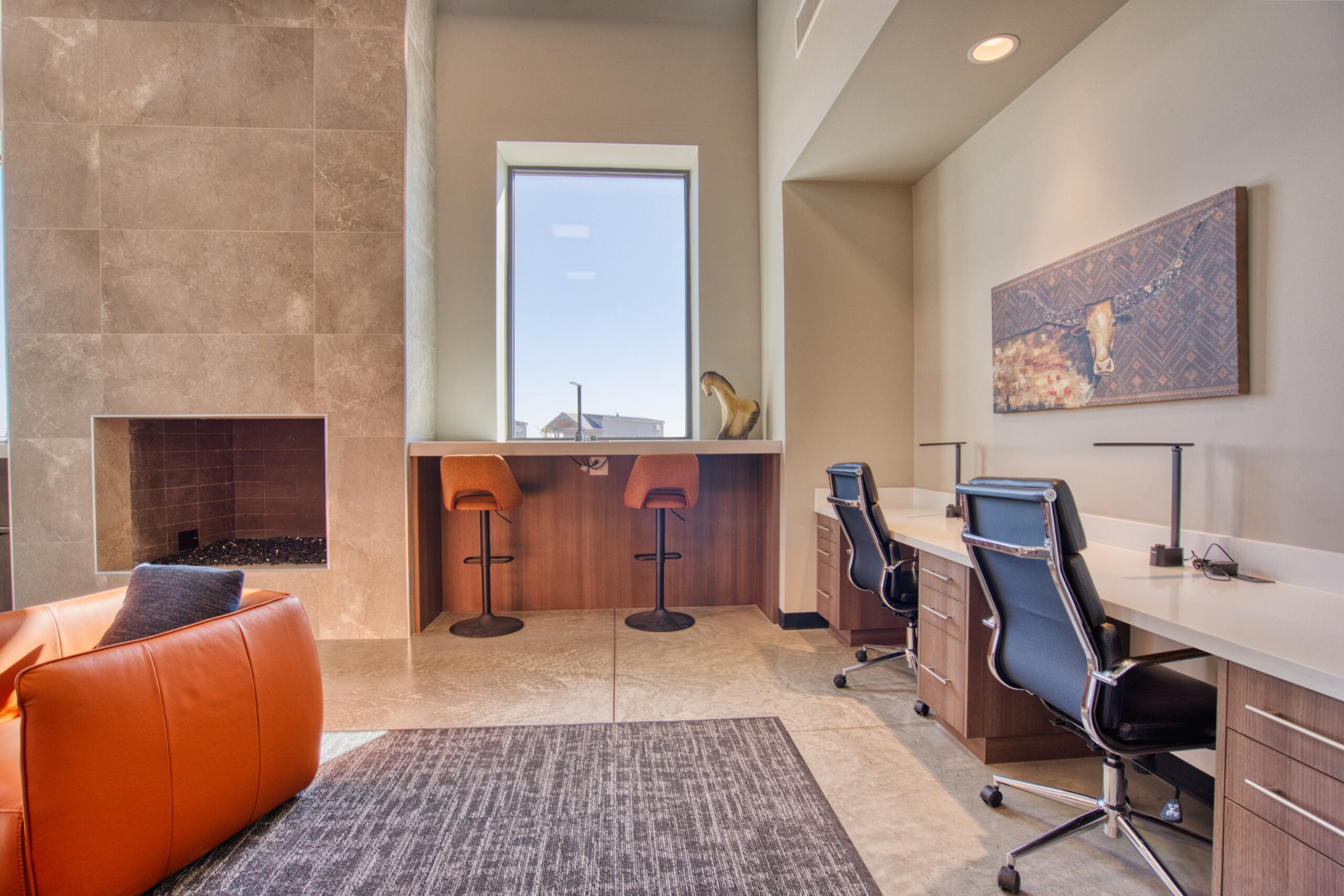 A modern workspace with two black office chairs at white desks, two orange barstools at a counter by a window, a large stone fireplace, an orange chair, and a framed artwork on the wall.