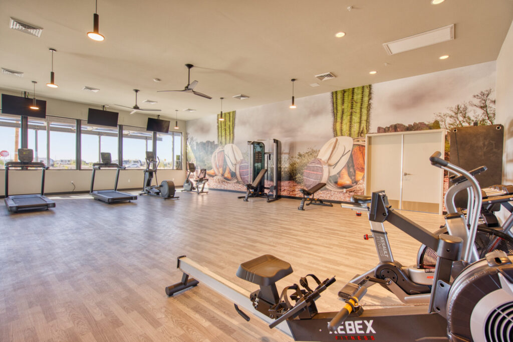 A spacious gym with treadmills, exercise bikes, rowing machines, and strength training equipment on wood flooring. A wall mural with desert plants and rocks decorates the room, and large windows let in natural light.