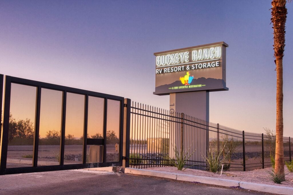 A large sign reading "Buckeye Ranch RV Resort & Storage" stands near a black metal fence and gate at sunset, with desert vegetation and palm trees visible in the background.
