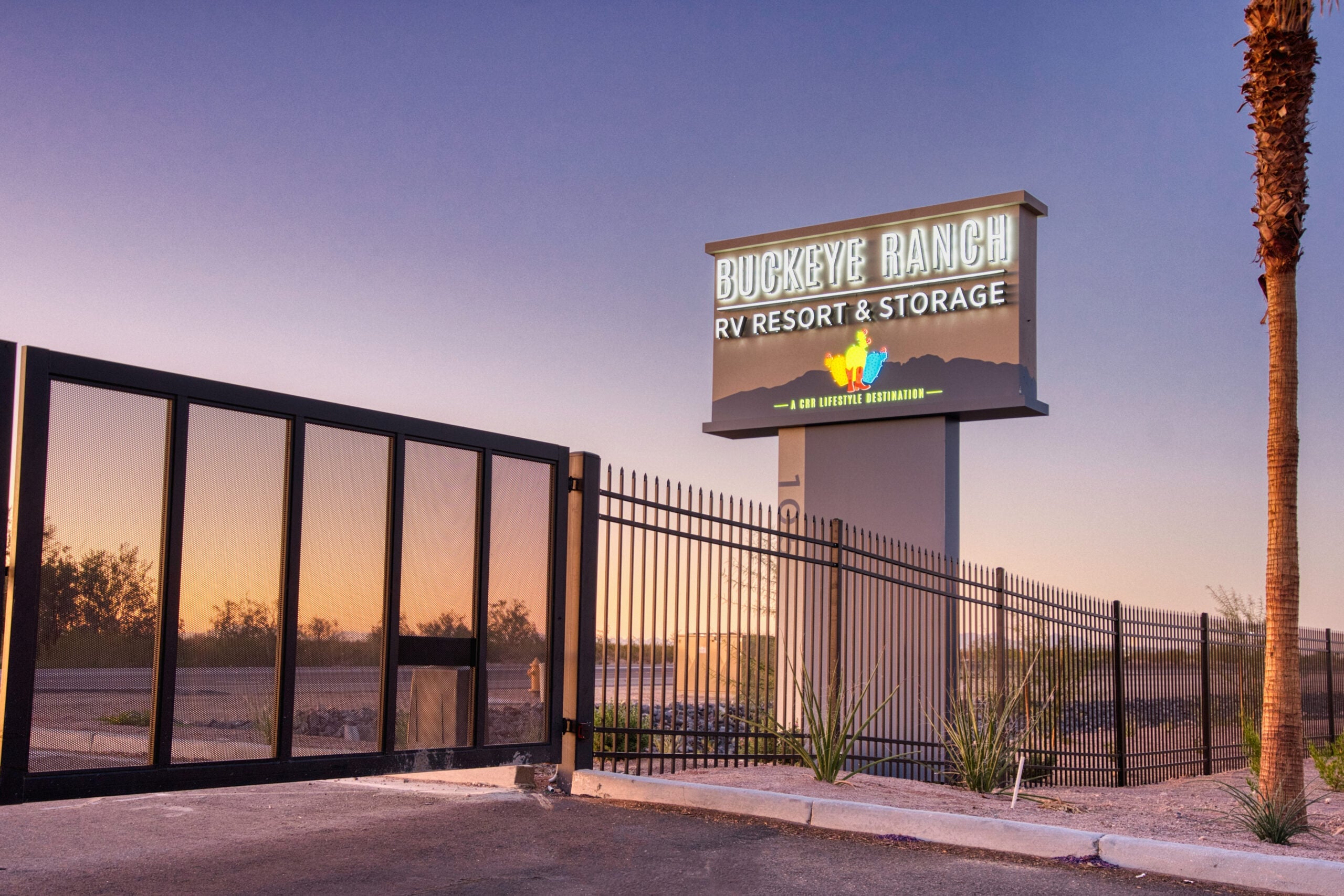 A large sign reading "Buckeye Ranch RV Resort & Storage" stands near a black metal fence and gate at sunset, with desert vegetation and palm trees visible in the background.