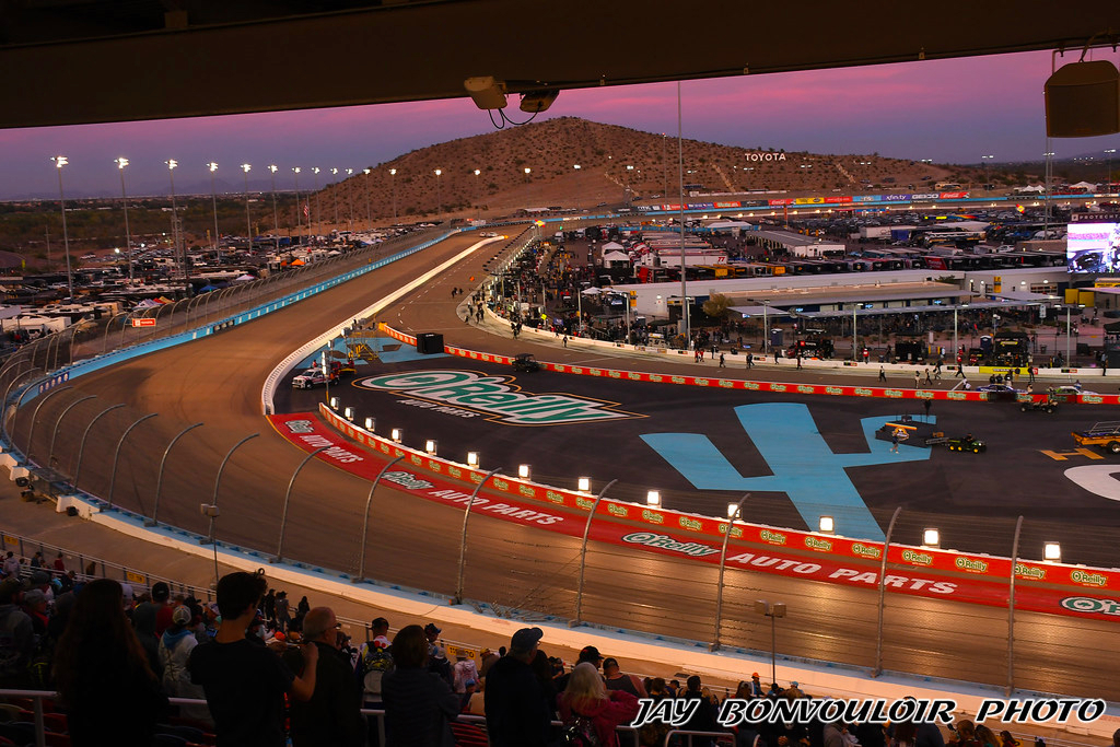 A crowd watches a sunset car race at Phoenix Raceway’s curved track lined with O'Reilly Auto Parts ads, with mountains in the background and brightly lit facilities nearby.