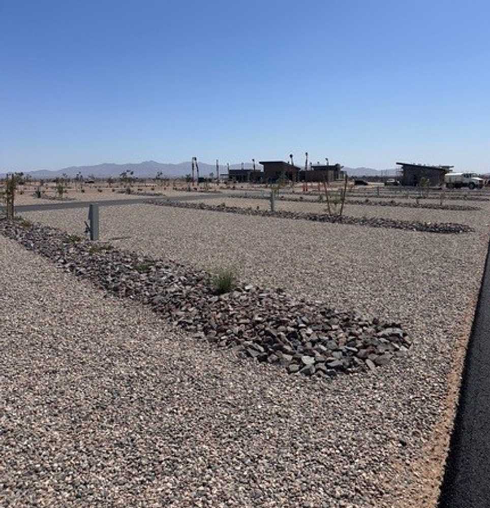 A gravel parking area in a dry, desert landscape with small shrubs, bordered by low rock dividers. Buildings and mountains are visible in the background under a clear blue sky.