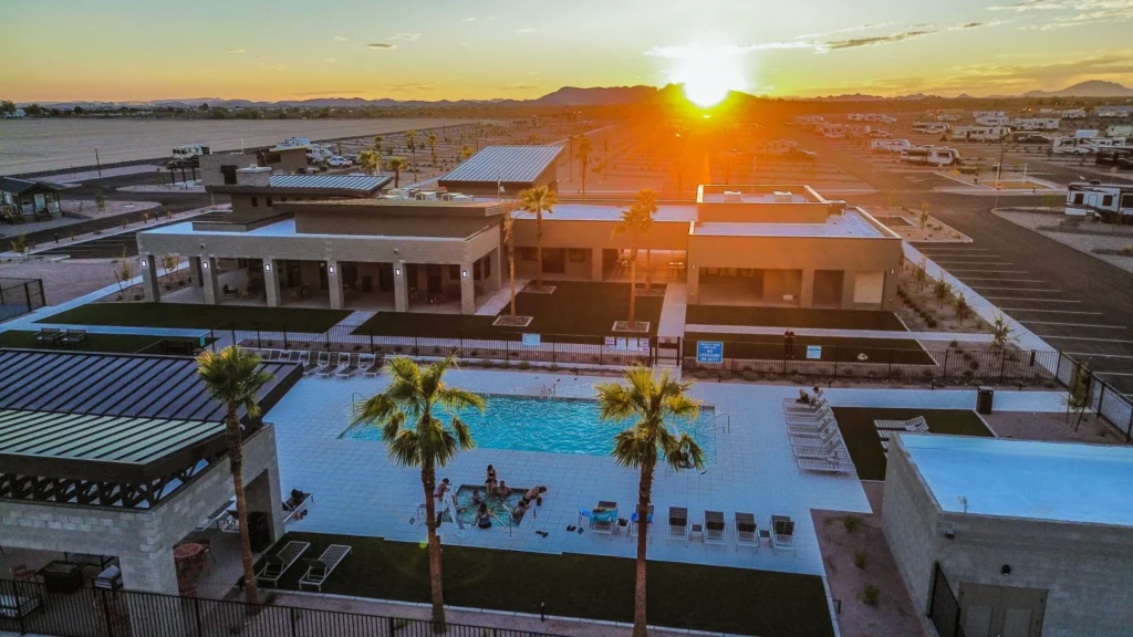 Aerial view of a modern recreational complex at sunset, featuring a swimming pool with people relaxing, palm trees, lounge chairs, and surrounding buildings, with RVs parked in the background.