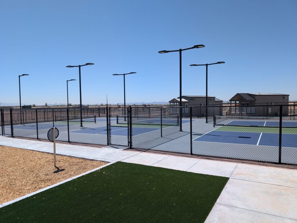 A fenced outdoor pickleball court complex with multiple courts, surrounded by streetlights and paved walkways, on a clear sunny day with houses and open land in the background.