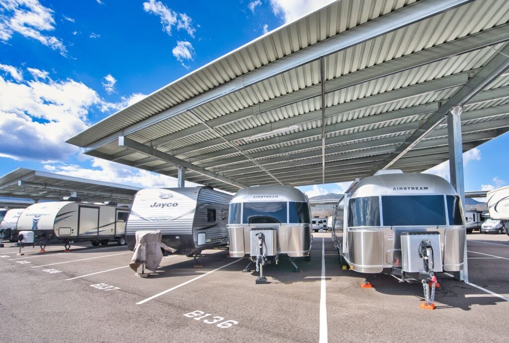 Rows of RVs and travel trailers, including Jayco and Airstream models, are parked under large metal canopies in a paved lot on a sunny day. Parking spaces are clearly marked with white lines and numbers.